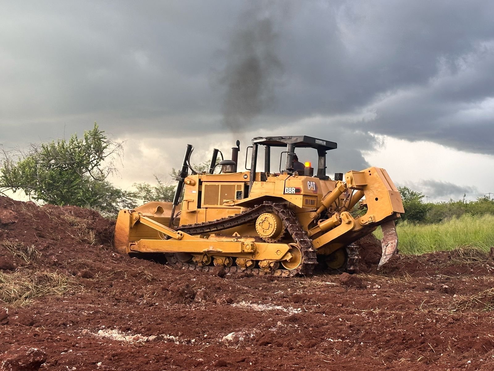 Yellow bulldozer clearing and flattening red soil under stormy sky with smoke in background