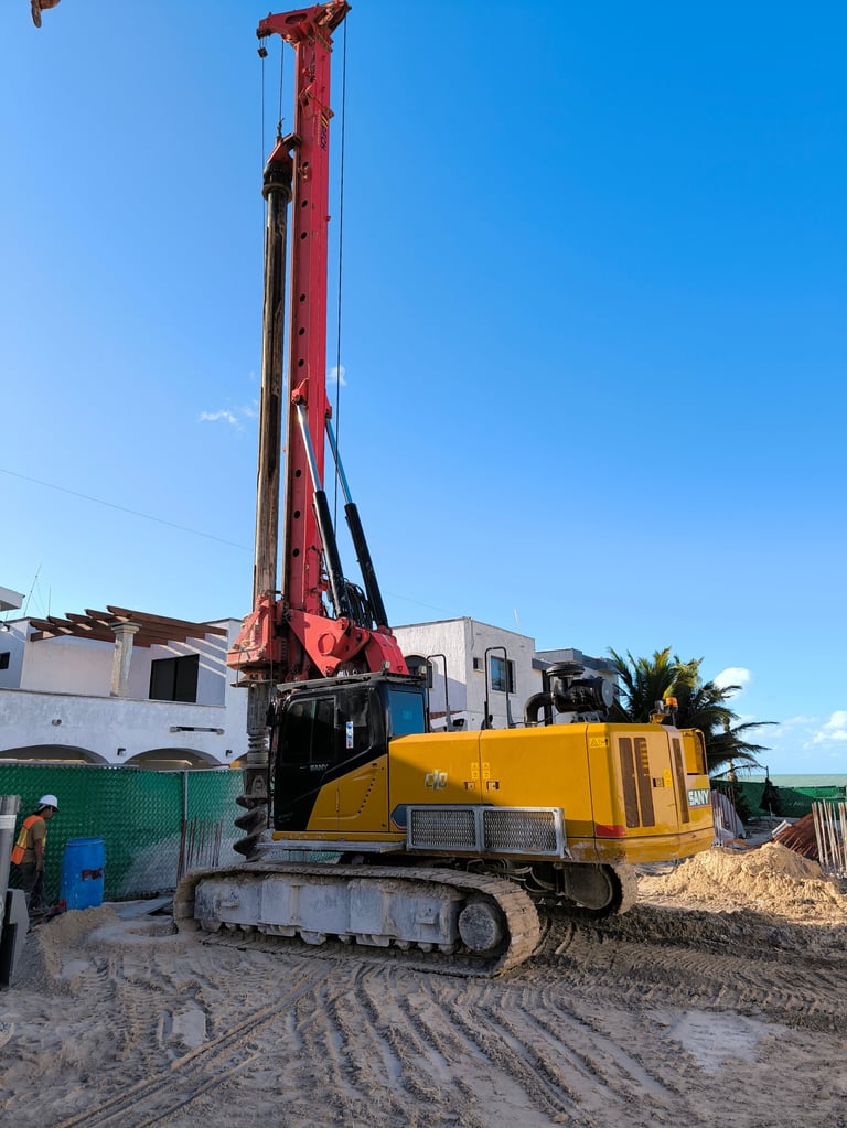 Yellow and red pile driver machine on a construction site with buildings and palm trees in the background under clear blue sky